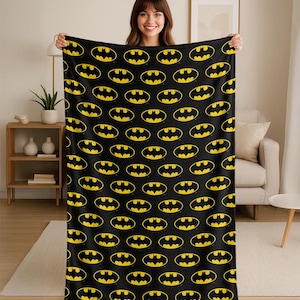 May include: Black blanket featuring a repeating pattern of yellow Batman logos. The logos are within an oval shape. The blanket is held up by a person. The blanket is a soft, cozy item.
