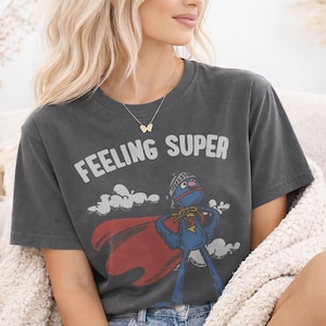 May include: Dark gray t-shirt with the words "FEELING SUPER" in white. The shirt features a cartoon character in a superhero costume with a red cape. The character is blue with a helmet and a "G" on its chest.