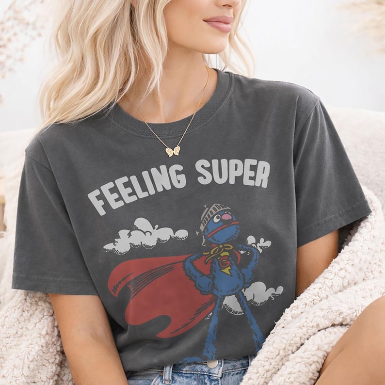 May include: Dark gray t-shirt with the words "FEELING SUPER" in white. The shirt features a cartoon character in a superhero costume with a red cape. The character is blue with a helmet and a "G" on its chest.