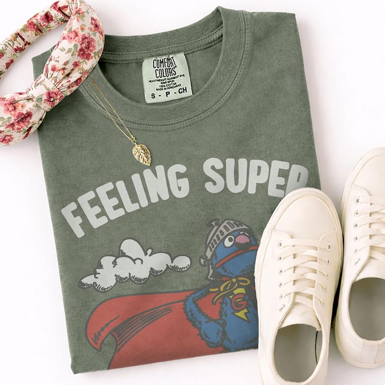 May include: An olive green t-shirt with the words "FEELING SUPER" printed across the front, along with a cartoon character in a superhero costume. A gold leaf pendant, floral headband, and white sneakers are also in the image.