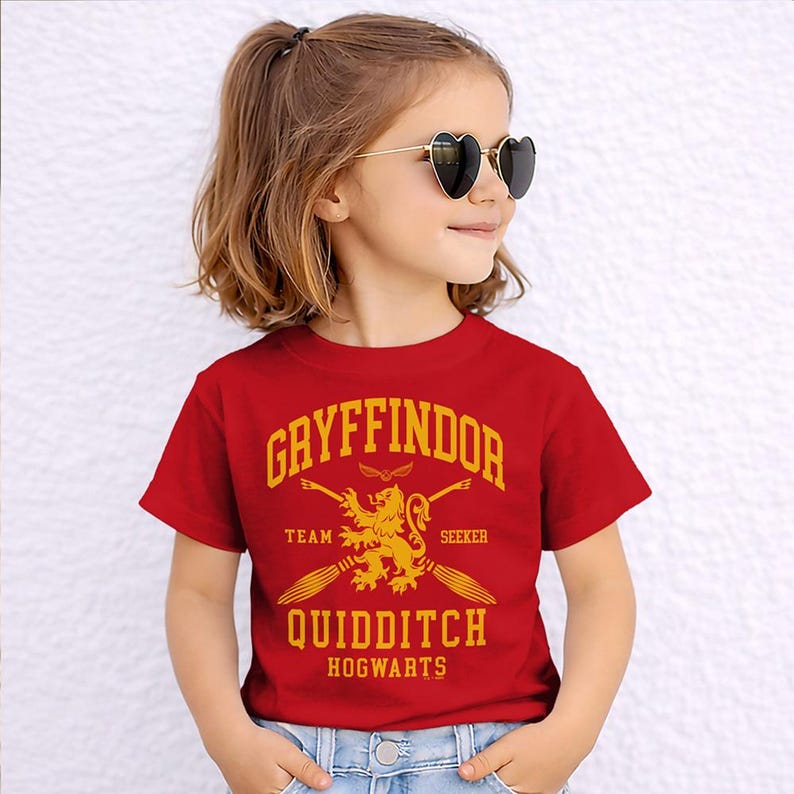 May include: A red t-shirt featuring the text "Gryffindor Team Seeker Quidditch Hogwarts" in gold lettering. The shirt has a graphic of a lion and two crossed brooms. The shirt is short-sleeved.