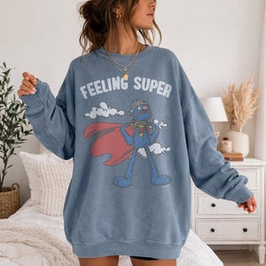 May include: A blue sweatshirt with the words "FEELING SUPER" in white. The graphic features a blue cartoon character with a red cape and a helmet. The sweatshirt is a solid blue color.