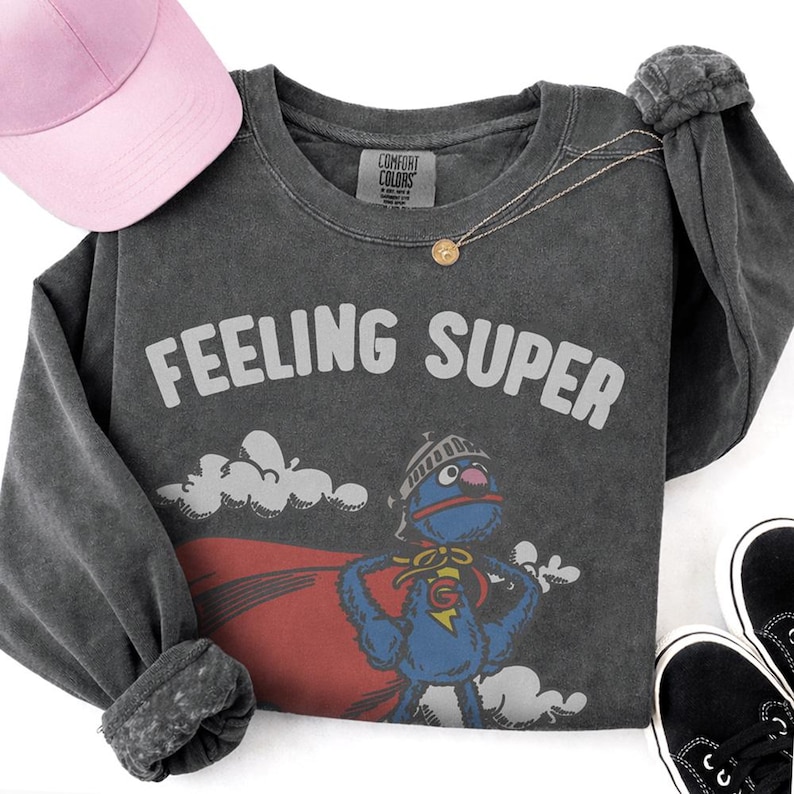 May include: Dark gray long-sleeve shirt with the text "FEELING SUPER" and a cartoon character graphic. A pink baseball cap, gold necklace, and black sneakers are also in the image.