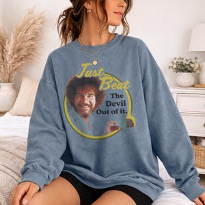 May include: A blue crewneck sweatshirt featuring a graphic print of Bob Ross holding a paintbrush inside a yellow circle. The text reads Just Beat The Devil Out of it in yellow and black lettering. The garment has long sleeves and a ribbed collar.