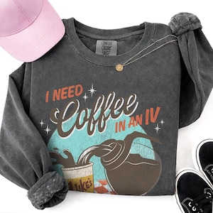 May include: Dark gray long-sleeve sweatshirt with the text "I NEED Coffee IN AN IV" in a retro design. A pink baseball cap, gold necklace, and black sneakers are also in the image. The sweatshirt has a distressed look.
