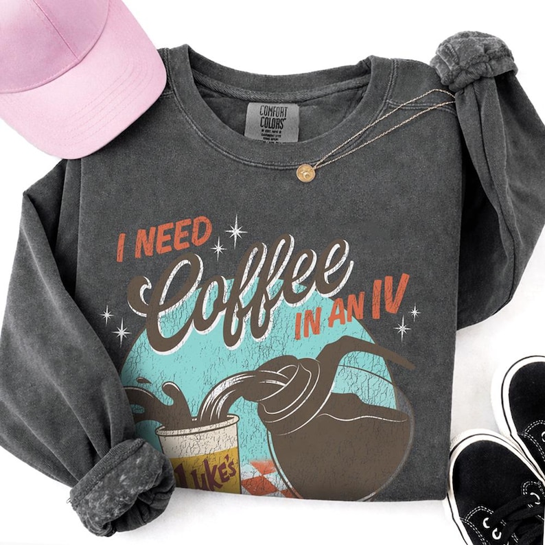 May include: Dark gray long-sleeve sweatshirt with the text "I NEED Coffee IN AN IV" in a retro design. A pink baseball cap, gold necklace, and black sneakers are also in the image. The sweatshirt has a distressed look.