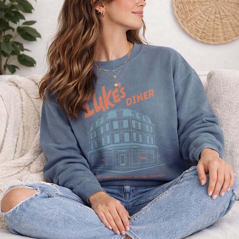 Op de afbeelding: Blauw sweatshirt met de oranje tekst "Luke's Diner" en een teal illustratie van een restaurant. Het sweatshirt toont ook de tekst "Breakfast Lunch & Dinner - NO CELL PHONES".