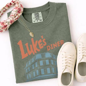 Op de afbeelding: Een olijfgroen t-shirt met de tekst "Luke's Diner" in oranje en een blauwe gebouwafbeelding. Een gouden blad hanger ketting, een bloemen haarband en witte sneakers staan ook in de afbeelding. Het shirt heeft een Comfort Colors label.