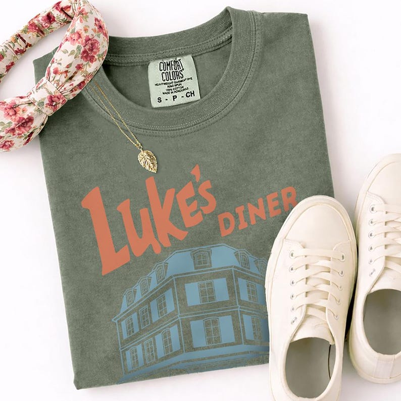 Op de afbeelding: Een olijfgroen t-shirt met de tekst "Luke's Diner" in oranje en een blauwe gebouwafbeelding. Een gouden blad hanger ketting, een bloemen haarband en witte sneakers staan ook in de afbeelding. Het shirt heeft een Comfort Colors label.