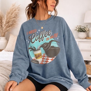 May include: Blue sweatshirt with the text "I NEED Coffee IN AN IV" and a graphic of coffee pouring from a carafe into a Luke's coffee cup. The graphic is set on a teal circle with a red and white checkered pattern.