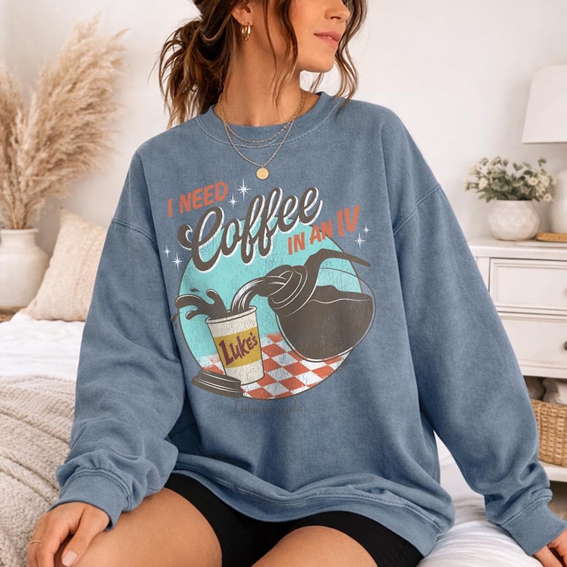 May include: Blue sweatshirt with the text "I NEED Coffee IN AN IV" and a graphic of coffee pouring from a carafe into a Luke's coffee cup. The graphic is set on a teal circle with a red and white checkered pattern.