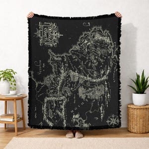 May include: Black throw blanket featuring a detailed map of Middle Earth in a cream color. The map includes intricate details of the land, with text and a compass rose. The blanket has a fringed edge.