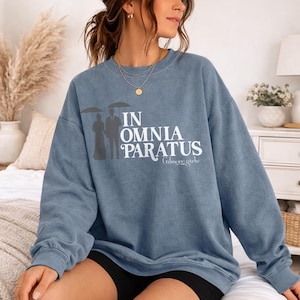May include: A blue-grey sweatshirt with the text "IN OMNIA PARATUS" and "Gilmore girls" printed in white. The design includes a silhouette of a couple under umbrellas. The sweatshirt is a comfortable, casual style.