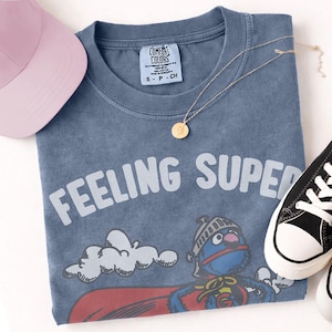 May include: A blue t-shirt with the words "FEELING SUPER" in white, featuring a cartoon character in a superhero costume. A pink baseball cap, gold necklace, and black and white sneakers are also in the image.