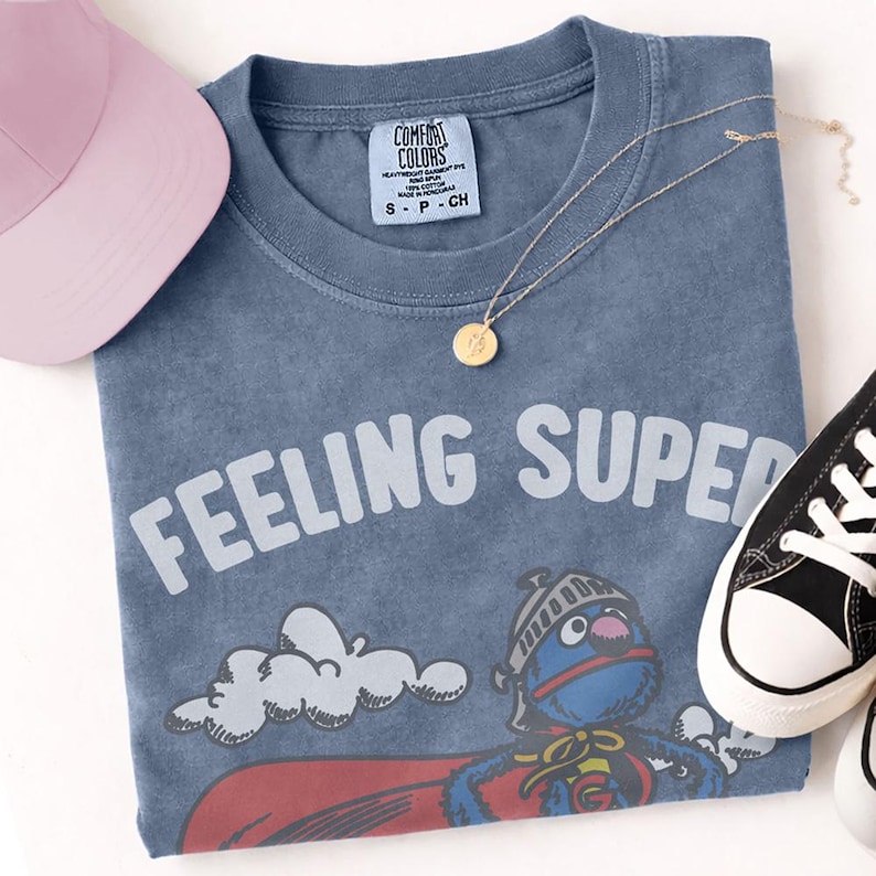 May include: A blue t-shirt with the words "FEELING SUPER" in white, featuring a cartoon character in a superhero costume. A pink baseball cap, gold necklace, and black and white sneakers are also in the image.