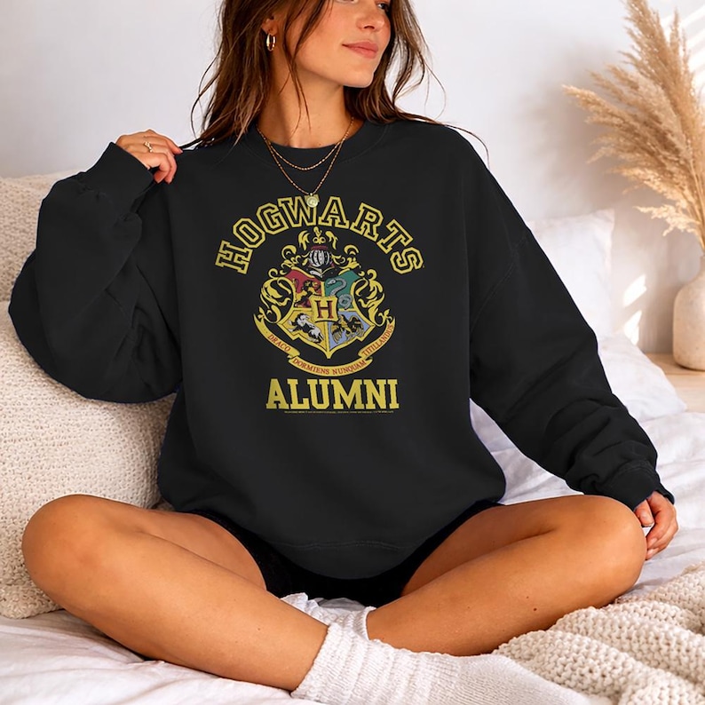 May include: Black sweatshirt featuring the Hogwarts crest in gold and the text "HOGWARTS ALUMNI". The crest includes the house colors and symbols. The sweatshirt is a comfortable, casual style.