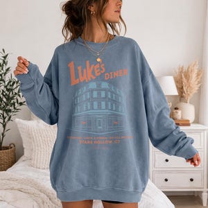 Op de afbeelding: Een blauwe sweatshirt met de oranje tekst "Luke's Diner" boven een teal illustratie van een diner. Onder de diner illustratie staat de tekst "BREAKFAST, LUNCH & DINNER - NO CELL PHONES" en "STARS HOLLOW, CT".