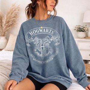 May include: A blue sweatshirt with the Hogwarts crest in white. The crest features a shield with the letter "H" and the words "Hogwarts" and Latin phrases. The sweatshirt has long sleeves and a crew neck.