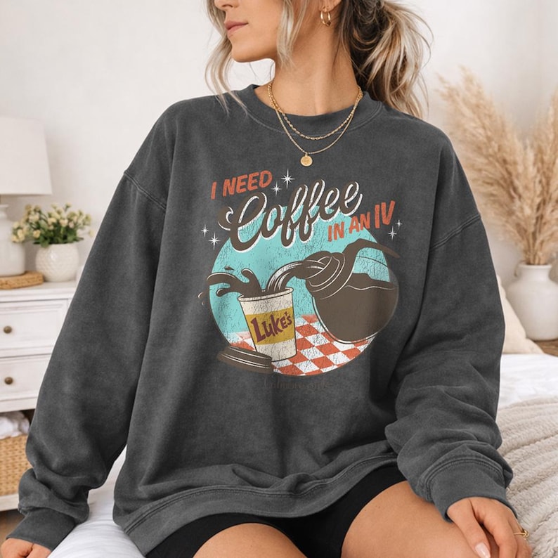 May include: Dark grey sweatshirt with the text "I NEED Coffee IN AN IV" above a graphic of coffee being poured into a cup. The cup is labeled "Luke's" and sits on a red and white checkered surface.