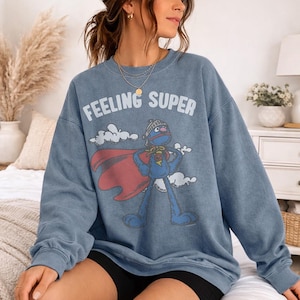 May include: A blue crewneck sweatshirt with the words "FEELING SUPER" in white. The graphic features a blue cartoon character wearing a cape and helmet. The character is standing in front of white clouds.
