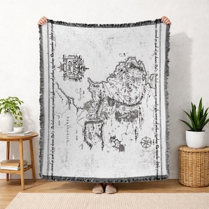 May include: A white and gray throw blanket featuring a detailed map design. The map includes intricate illustrations of landscapes, text, and a compass rose. The blanket has a fringed edge and is held up by a person.
