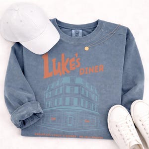Op de afbeelding: Een blauwe sweatshirt met de tekst "Luke's Diner" in oranje en een gebouwafbeelding. Een witte baseballpet, een gouden ketting en witte sneakers staan ook op de foto. De tekst onderaan luidt "Breakfast, Lunch, & Dinner - No Cell Phones."