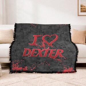 May include: A black throw blanket with the words "I ❤️ DEXTER" in red, displayed on a white couch. The blanket has a distressed design with red splatter accents and a fringed edge. The couch is in a living room setting.