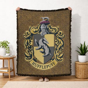 May include: A brown and gold Hufflepuff throw blanket. The blanket features the Hufflepuff crest, including a badger, shield, and the word "Hufflepuff". The blanket has a patterned background and a fringed edge.
