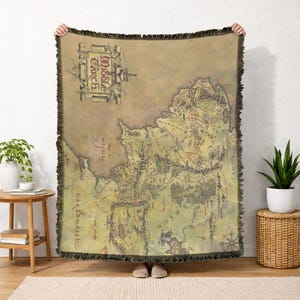 May include: A tan throw blanket featuring a detailed map of Middle Earth. The map is in shades of green, brown, and red, with the words "Middle Earth" in a decorative banner. The blanket has a fringed edge.