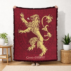 May include: A burgundy throw blanket featuring a golden lion rampant emblem, with the words "GAME OF THRONES" at the bottom. The blanket has a fringed edge and a detailed pattern in the background.