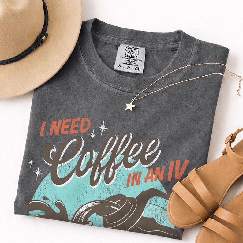 May include: A charcoal gray t-shirt with the phrase "I NEED Coffee IN AN IV" in white and orange lettering. The shirt features a coffee cup graphic with blue and brown accents. A gold star pendant necklace, a tan hat, and brown sandals are also in the image.
