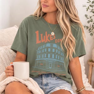 Op de afbeelding: Olijfgroen t-shirt met de tekst "Luke's Diner" in oranje en een blauw-bruine illustratie van een restaurant. Het shirt bevat ook de tekst "Breakfast, Lunch, & Dinner, No Cell Phones".