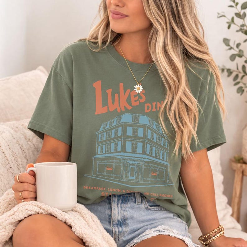 Op de afbeelding: Olijfgroen t-shirt met de tekst "Luke's Diner" in oranje en een blauw-bruine illustratie van een restaurant. Het shirt bevat ook de tekst "Breakfast, Lunch, & Dinner, No Cell Phones".