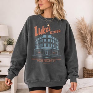 Op de afbeelding: Donkergrijze sweatshirt met de tekst "Luke's Diner" in oranje en een blauw-oranje illustratie van een eetcaf&eacute;. De tekst "BREAKFAST, LUNCH, & DINNER - NO CELL PHONES STARS HOLLOW, CT" is onder de afbeelding gedrukt.