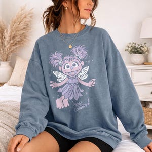May include: Blue-grey sweatshirt featuring a cartoon character with purple hair and wings. The character is wearing a purple dress and has the text "Abby Cadabby!" below. The sweatshirt is a crew neck style.