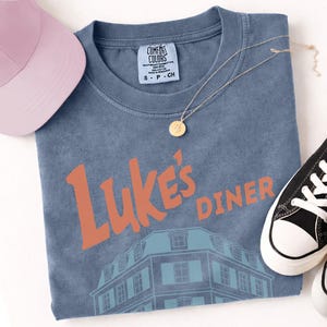 Op de afbeelding: Een blauw Comfort Colors T-shirt met de tekst "Luke's Diner" in oranje en een gebouwafbeelding. Een roze baseballpet, een gouden ketting en zwart-witte sneakers staan ook op de foto.