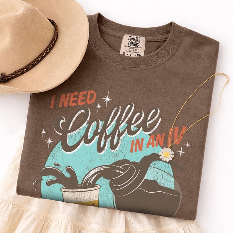 May include: A brown t-shirt with the text "I NEED Coffee IN AN IV" in white, red, and blue. The graphic shows coffee pouring into a cup. A tan hat with a brown band and a gold necklace with a daisy pendant are also visible.