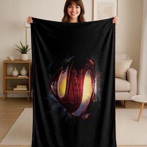 Puede incluir: Manta de forro polar negra con el logotipo de Superman desgastado en rojo, amarillo y azul. La manta est&aacute; sostenida por una persona, mostrando su tama&ntilde;o y dise&ntilde;o. Un art&iacute;culo de decoraci&oacute;n del hogar acogedor.