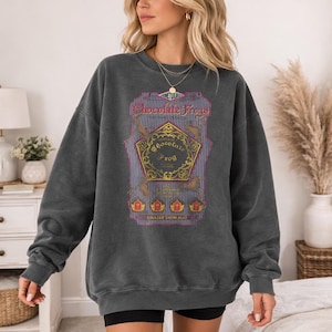 May include: Dark gray sweatshirt with a graphic featuring the text "Chocolate Frogs" and a gold and purple design. The graphic includes images of frogs and the text "Collect Them All".