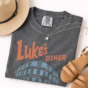 Op de afbeelding: Een antracietgrijs t-shirt met de woorden "Luke's Diner" in oranje en een blauwe gebouwafbeelding. Een gouden sterhangerketting, bruinleren sandalen en een bruine hoed staan ook op de foto.