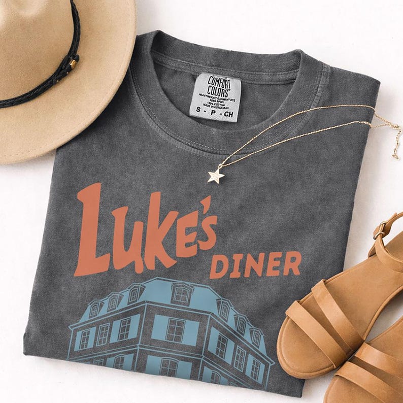 Op de afbeelding: Een antracietgrijs t-shirt met de woorden "Luke's Diner" in oranje en een blauwe gebouwafbeelding. Een gouden sterhangerketting, bruinleren sandalen en een bruine hoed staan ook op de foto.