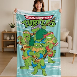 May include: A light blue blanket featuring the Teenage Mutant Ninja Turtles. The blanket has a teal background with white brushstroke accents. The Turtles are depicted in their signature colors: red, orange, blue, and purple. The blanket has the words "Teenage Mutant Ninja Turtles" in a red and green font.