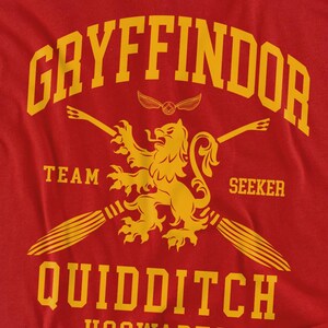 May include: Red t-shirt with a yellow Gryffindor Quidditch design. The design includes the word "Gryffindor" at the top, a lion emblem, the words "Team Seeker", and the word "Quidditch" at the bottom. The shirt is made of a soft material.