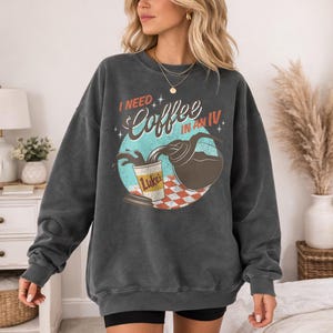 May include: Dark gray sweatshirt with the text "I NEED Coffee IN AN IV" and a graphic of coffee being poured into a cup. The cup has the name "Luke's" on it. The graphic is set against a teal and red background.