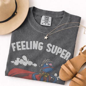 May include: A charcoal gray t-shirt with the words "FEELING SUPER" in white. The shirt features a cartoon character in a superhero costume. A gold star pendant necklace, tan sandals, and a tan hat with a black band are also in the image.