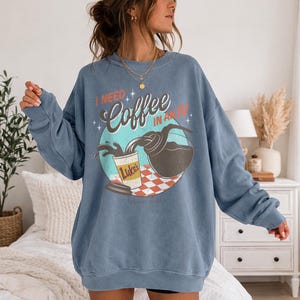 May include: Blue crewneck sweatshirt with a graphic that reads "I NEED Coffee IN AN IV." The graphic features a coffee pot pouring coffee into a Luke's coffee cup. The design is set against a teal and red checkered background.