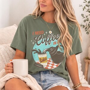 May include: Olive green t-shirt with the text "I NEED Coffee IN A..." and a graphic of coffee being poured into a cup. The cup is labeled "Luke's". The shirt is worn by a person.
