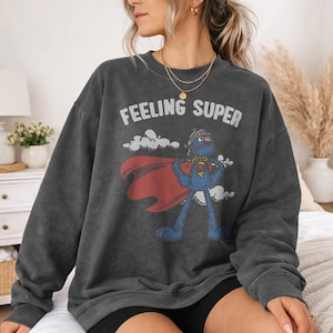 May include: Dark gray sweatshirt with the words "FEELING SUPER" above a cartoon character in a superhero costume. The character is blue with a red cape and a helmet. The sweatshirt is a vintage style.