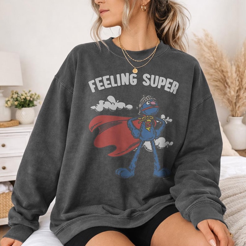 May include: Dark gray sweatshirt with the words "FEELING SUPER" above a cartoon character in a superhero costume. The character is blue with a red cape and a helmet. The sweatshirt is a vintage style.