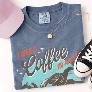 May include: A blue Comfort Colors t-shirt with the text "I NEED Coffee IN AN IV". A pink baseball cap, gold necklace, and black and white sneakers are also in the image. The shirt has a vintage, distressed look.